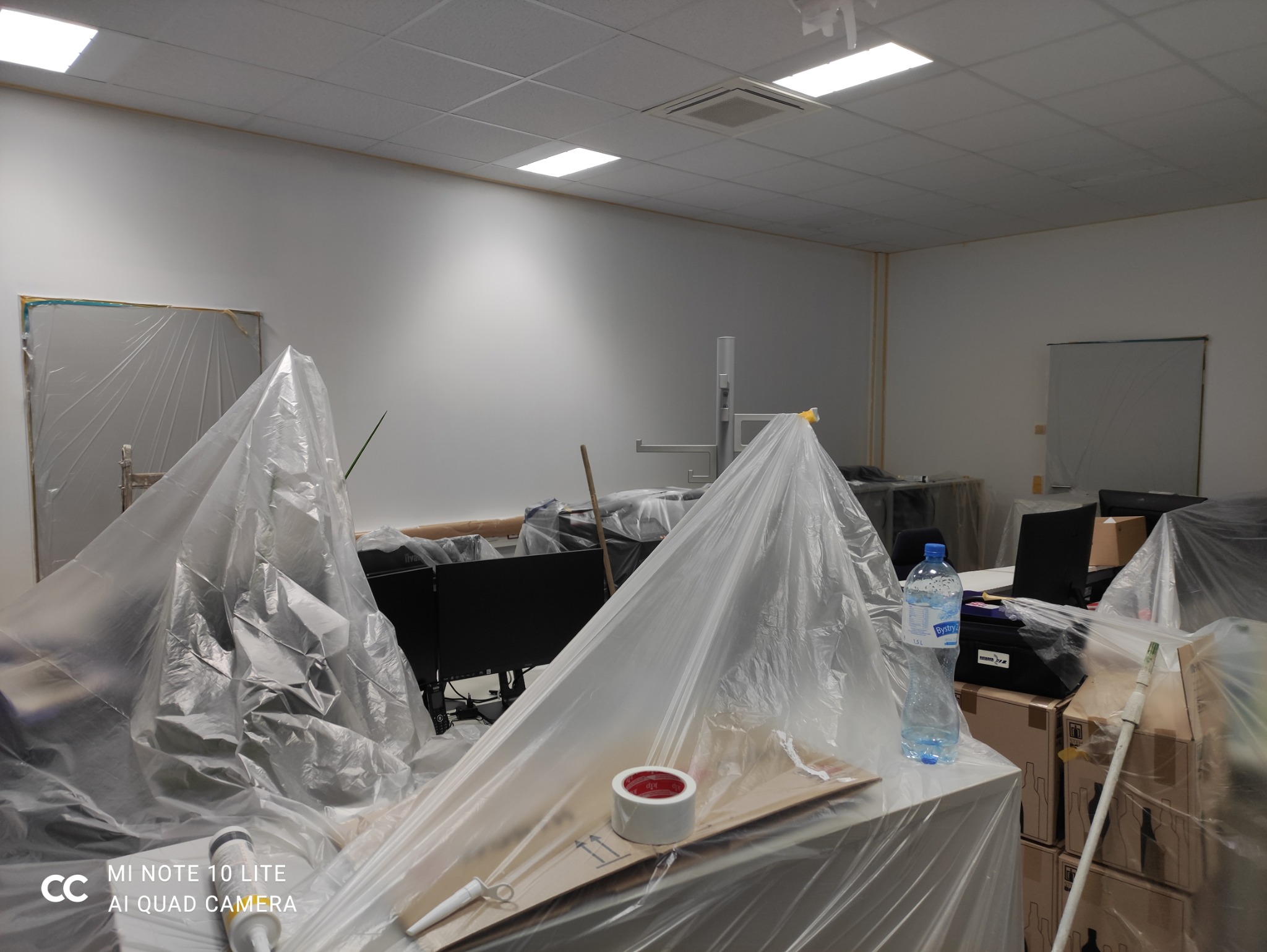 Przygotowanie biura do malowania: meble i sprzęt zabezpieczone folią ochronną, widoczne narzędzia malarskie. Oświetlenie sufitowe.
