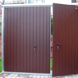 Nowoczesna, brązowa brama wjazdowa z metalową ramą, częściowo otwarta, z widocznymi zamkami i kluczami, na tle kostki brukowej i zieleni.