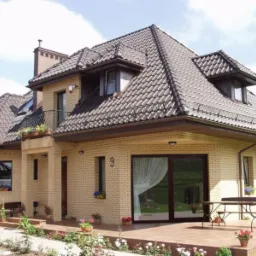 Elewacja dwupiętrowego domu z beżowej cegły klinkierowej, z brązowym dachem z dachówki ceramicznej, balkonem z kutą balustradą i tarasem z przesuwnymi drzwiami balkonowymi, numer domu '9' widoczny...