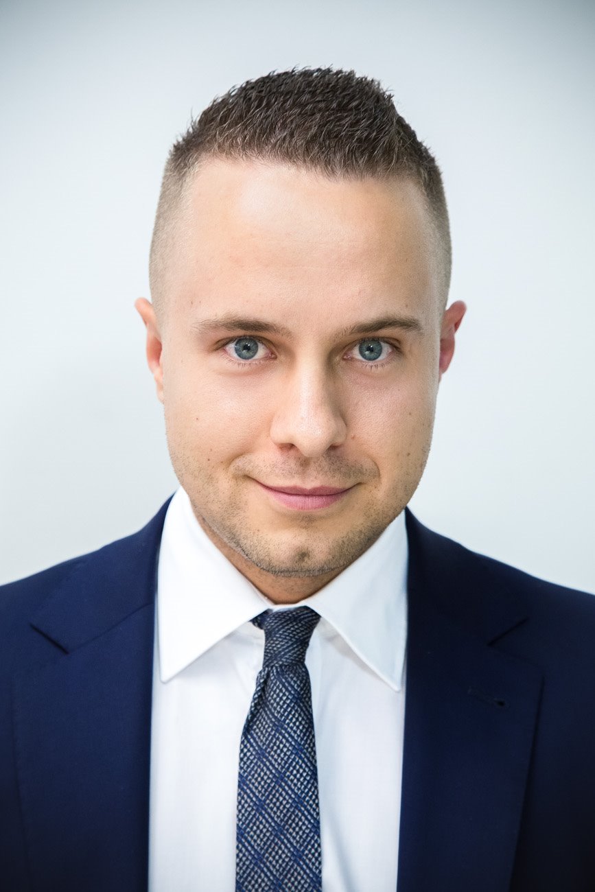 Portret uśmiechniętego mężczyzny w garniturze z niebieskimi oczami i krótką fryzurą na jasnym tle.