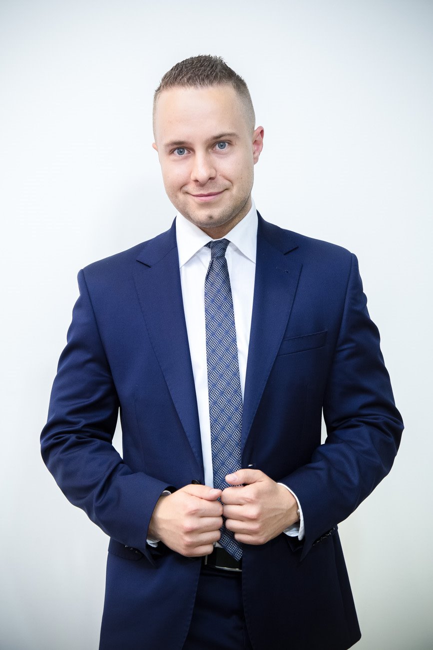 Uśmiechnięty mężczyzna w granatowym garniturze i wzorzystym krawacie, z rękami splecionymi na wysokości brzucha, pozuje na jasnym tle.