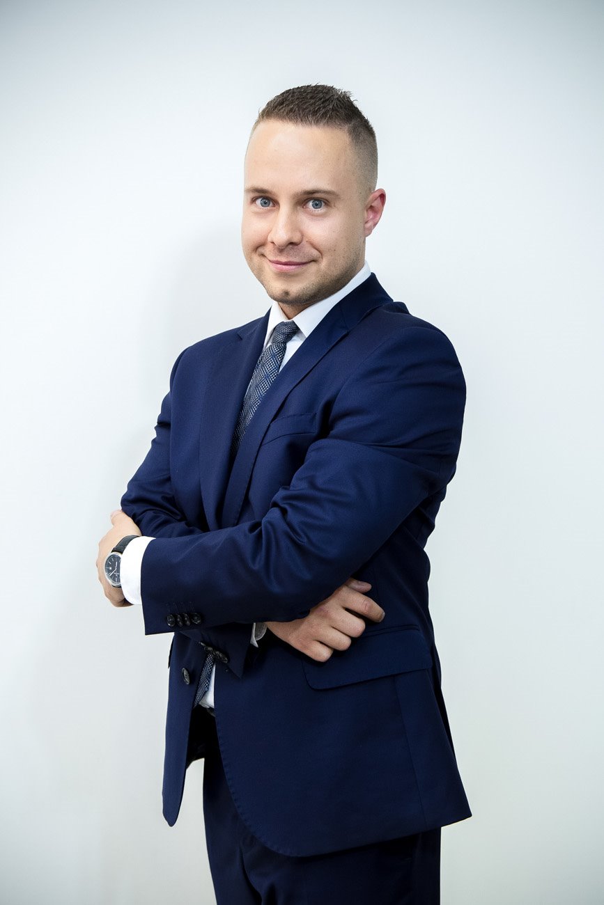 Portret uśmiechniętego mężczyzny w granatowym garniturze, stojącego na tle białej ściany z założonymi rękami i widocznym zegarkiem na lewym nadgarstku.