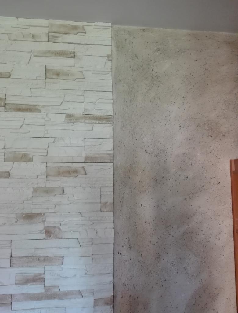 Fragment ściany z dekoracyjną okładziną imitującą kamień, sąsiadującej z fragmentem ściany pokrytej szarą farbą strukturalną z ciemnymi punktami, widoczna krawędź drewnianej framugi drzwi.