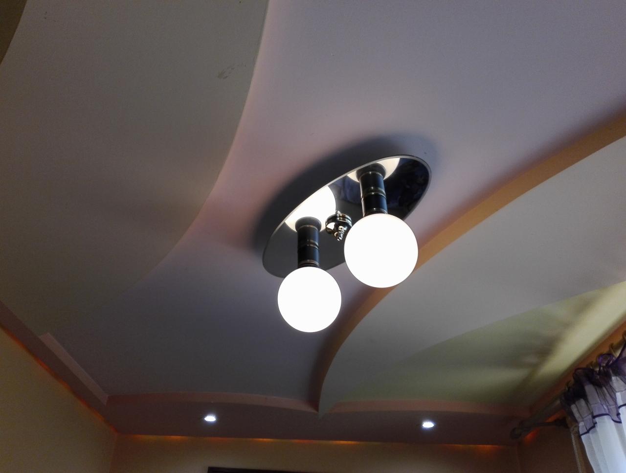 Kremowy sufit wielopoziomowy z dekoracyjnymi wgłębieniami i nowoczesnym oświetleniem punktowym oraz lampą wiszącą z dwiema kulistymi żarówkami.