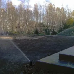 Automatyczne nawadnianie świeżo przygotowanej gleby pod trawnik, z widocznymi zraszaczami i brzegiem tarasu, w tle las brzozowy.