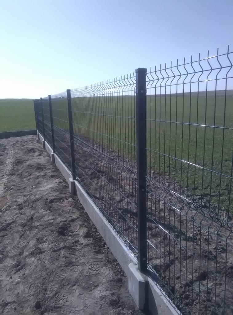 Nowoczesne ogrodzenie panelowe z podmurówką betonową na granicy działki z polem uprawnym, widoczne ślady prac ziemnych.
