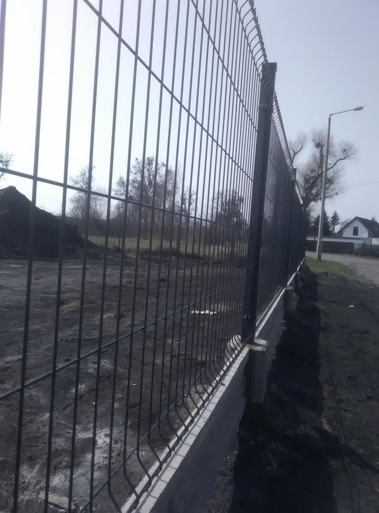 Nowoczesne ogrodzenie panelowe z podmurówką betonową, widok z boku, z czarną ziemią i fragmentem ulicy w tle.