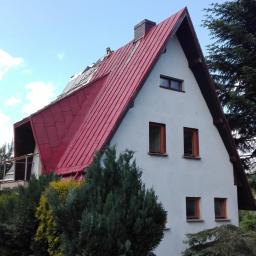 Normal-rembud Wojciech Rutkowski - Dwuspadowy dom z czerwonym dachem z blachy, częściowo zdemontowanym, widok z poziomu ogrodu z zielenią i drzewami.