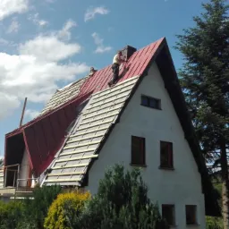 Prace dekarskie w Goleszowie: montaż czerwonego pokrycia na dachu dwuspadowym domu z białymi ścianami, widoczne deski podkładowe i dekarz pracujący na górze.