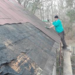 Normal-rembud Wojciech Rutkowski - Pracownik w zielonej bluzie na dachu z częściowo zdjętym, starym pokryciem, montujący nowe panele blaszane, widok z dołu na tle drzew.