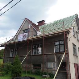 Normal-rembud Wojciech Rutkowski - Dom jednorodzinny w trakcie prac dekarskich, widoczne rusztowanie, częściowo zdjęte stare pokrycie i przygotowane podłoże pod nowe, baner reklamowy firmy dekarskiej na rusztowaniu.