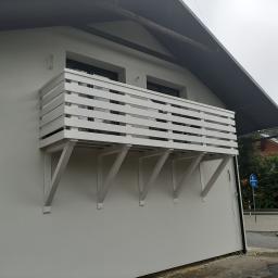 Normal-rembud Wojciech Rutkowski - Biały, drewniany balkon z poziomymi balustradami, wsparty na trzech wspornikach, zamontowany na ścianie budynku z szarą elewacją, widok z dołu na tle pochmurnego nieba.