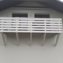 Normal-rembud Wojciech Rutkowski - Biały balkon z drewnianą balustradą i wspornikami, zamontowany na elewacji budynku z widocznymi oknami w tle.