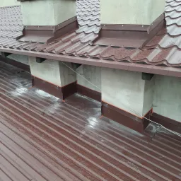 Brązowy blaszany dach z kominami obrobionymi blachą tego samego koloru, widoczne ślady wody, elementy mocujące i uszczelnienia.