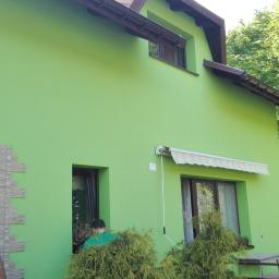 Normal-rembud Wojciech Rutkowski - Jasnozielony dom z brązowymi oknami i dachem, częściowo wykończony kamieniem elewacyjnym przy wejściu, z rozwiniętą markizą nad oknem i osobami w pobliżu.