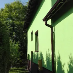 Jasnozielona elewacja domu jednorodzinnego w Goleszowie, z brązowymi rynnami i podmurówką, widziana z boku na tle zieleni drzew.