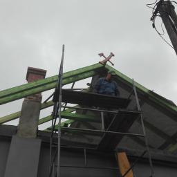 Normal-rembud Wojciech Rutkowski - Pracownik na rusztowaniu montuje elementy konstrukcyjne dachu z zielonego drewna, widoczny komin z cegły i ozdobny element na szczycie dachu w pochmurny dzień.