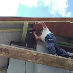 Normal-rembud Wojciech Rutkowski - Monter na rusztowaniu mocuje drewnianą podbitkę dachową za pomocą narzędzia pneumatycznego, widoczne elementy konstrukcji dachu, rynna i fragment elewacji budynku.