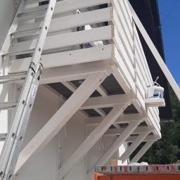 Normal-rembud Wojciech Rutkowski - Biała elewacja balkonu z drewnianą balustradą w trakcie malowania, widoczna drabina oparta o ścianę i wiadro z białą farbą na półce rusztowania.