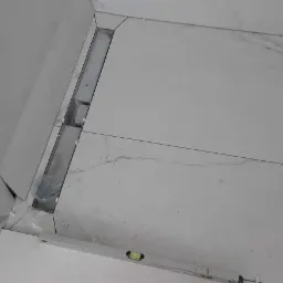 Białe płytki podłogowe z marmurowym wzorem w trakcie układania, widoczny liniowy odpływ prysznicowy i poziomica.