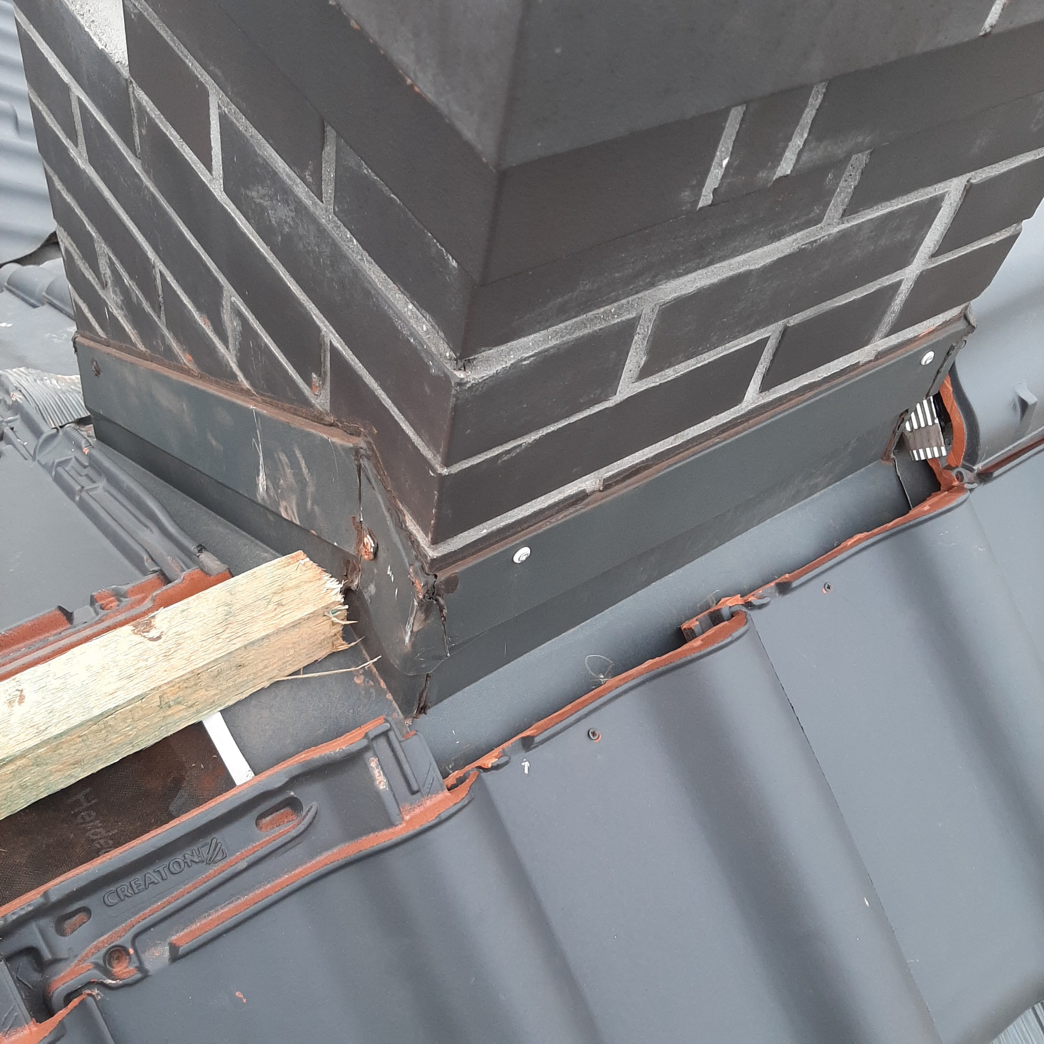 Ciemny komin murowany z cegły na dachu krytym szarą dachówką ceramiczną CREATON, widoczne elementy obróbek blacharskich i fragment deski.
