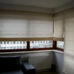 Beżowe rolety rzymskie zamontowane na dwóch oknach w rogu pokoju, widoczny fragment czarnej sofy i kaloryfer.