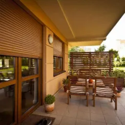 Brązowe rolety zewnętrzne częściowo opuszczone na oknach tarasu, drewniane meble ogrodowe i dekoracyjna przesłona w tle.