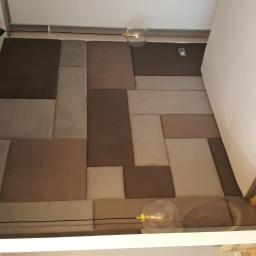 Meb-lokum - Panel tapicerowany z kwadratowych i prostokątnych elementów w odcieniach brązu i szarości, tworzący zagłówek łóżka, oświetlony nowoczesnymi lampami z przezroczystymi kloszami.