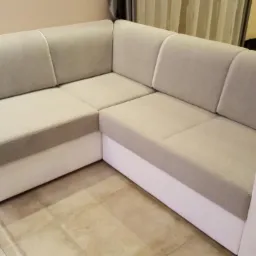 Narożnik z jasnoszarą tapicerką i białym wykończeniem, widoczny fragment szklanego stolika kawowego z metalowymi elementami.