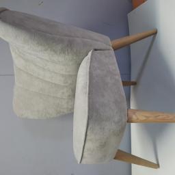 Meb-lokum - Jasnoszary fotel z drewnianymi nogami, widoczne przeszycia na oparciu, ustawiony na tle ściany i krawędzi pomalowanej na pomarańczowo z czarnymi plamami.