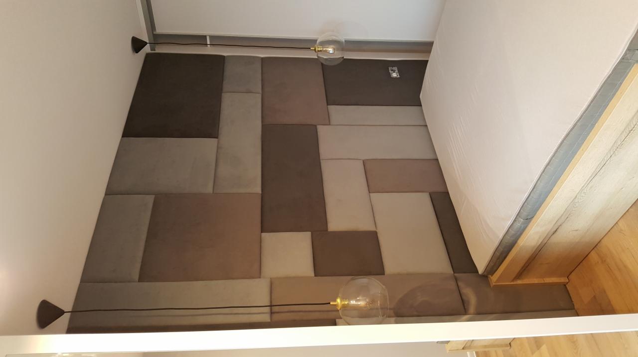 Panel tapicerowany z kwadratowych i prostokątnych elementów w odcieniach brązu i szarości, tworzący zagłówek łóżka, oświetlony nowoczesnymi lampami z przezroczystymi kloszami.