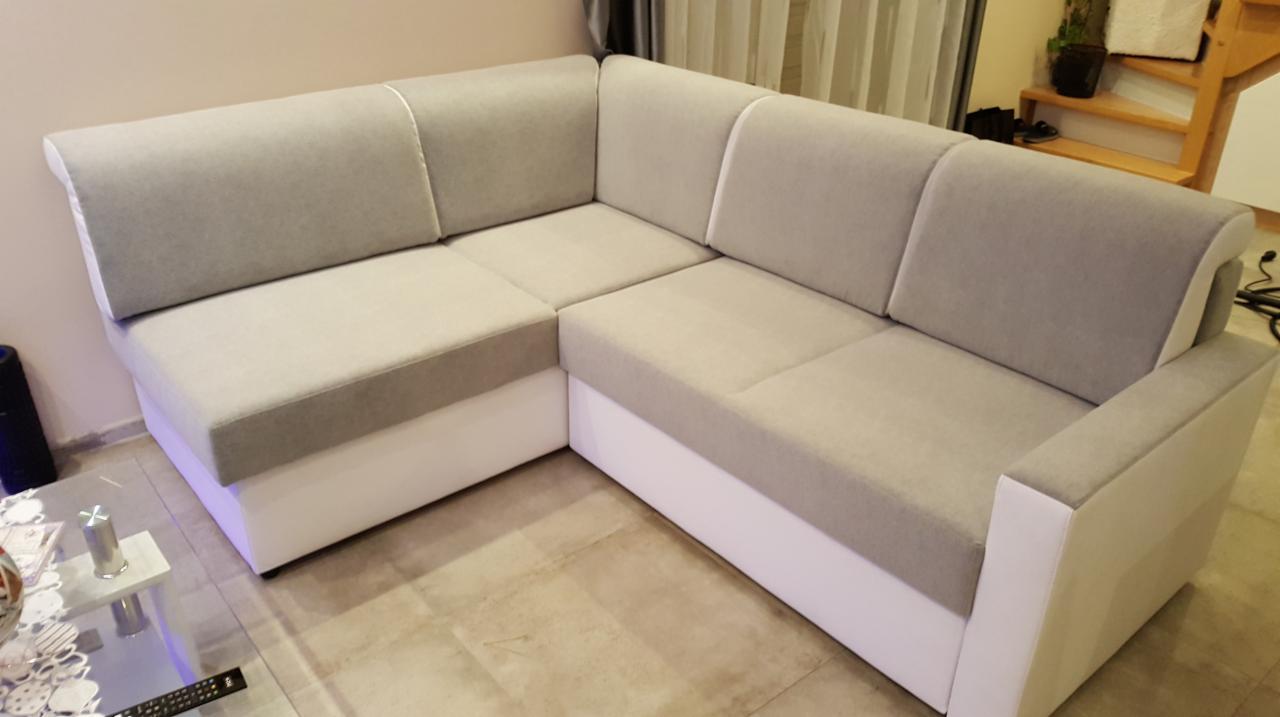 Narożnik z jasnoszarą tapicerką i białym wykończeniem, widoczny fragment szklanego stolika kawowego z metalowymi elementami.