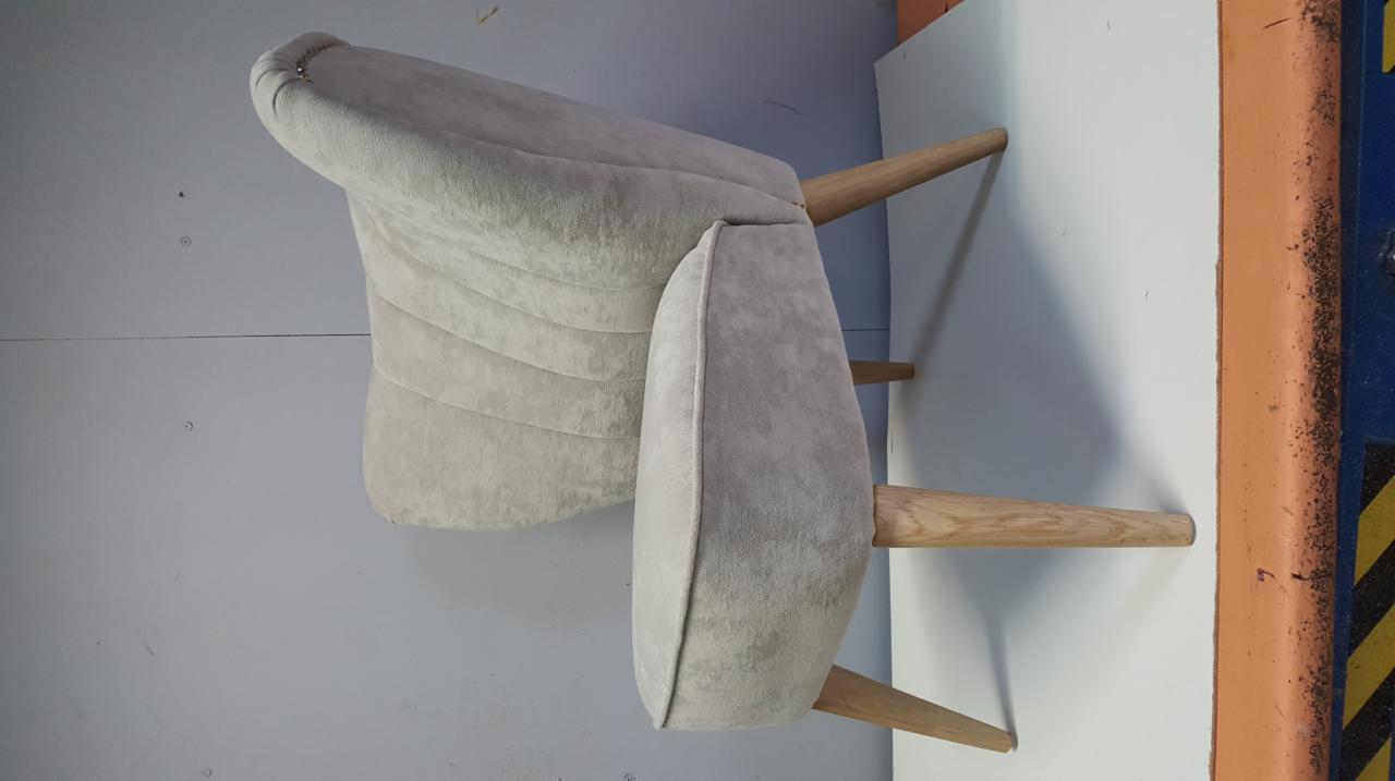 Jasnoszary fotel z drewnianymi nogami, widoczne przeszycia na oparciu, ustawiony na tle ściany i krawędzi pomalowanej na pomarańczowo z czarnymi plamami.