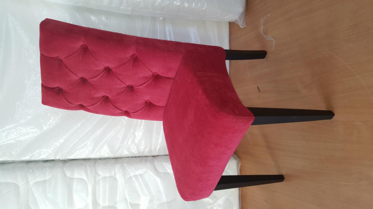 Czerwone, tapicerowane wezgłowie łóżka z pikowaniem, oparte o materac w folii ochronnej, widoczne czarne nóżki.