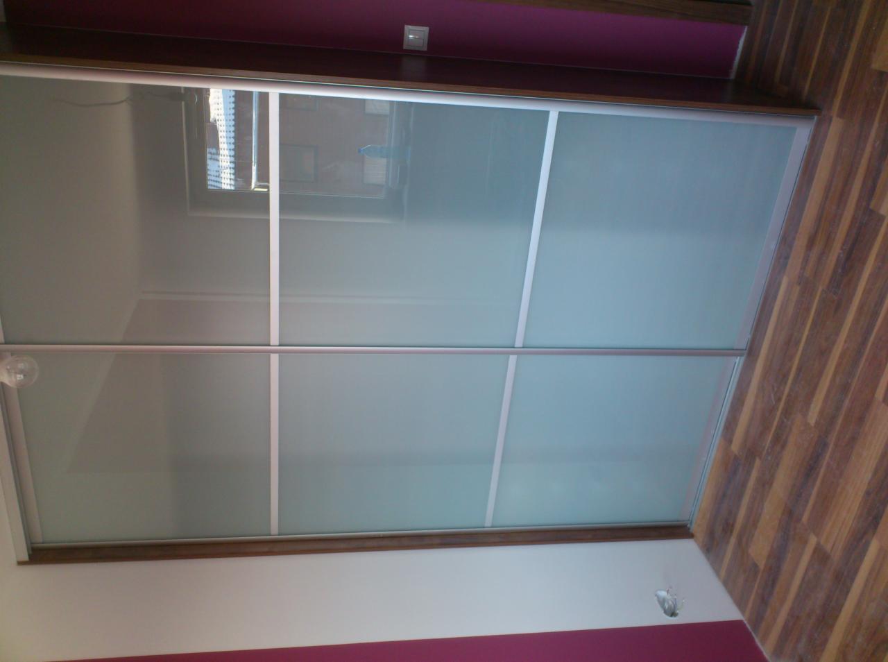 Wbudowana szafa z przesuwnymi drzwiami z matowego szkła i aluminiowymi ramami, widoczna z perspektywy pod kątem, z fragmentem drewnianej podłogi i ściany w dwóch odcieniach.