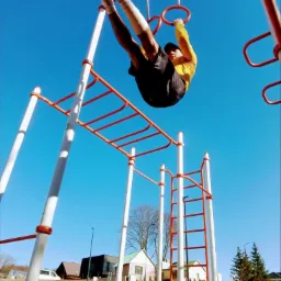 Mężczyzna w żółtej koszulce i czarnych spodenkach wykonuje ćwiczenie na drążku na placu do street workoutu, trzymając się za kółka, z nogami uniesionymi do góry na tle błękitnego nieba.