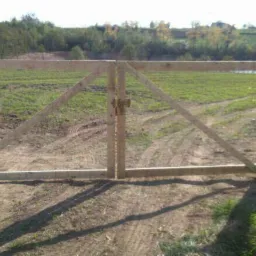 Drewniana dwuskrzydłowa brama polna z prostym zamkiem, osadzona w drewnianych słupkach, z siatką ogrodzeniową w tle i widocznymi śladami opon na ziemi.