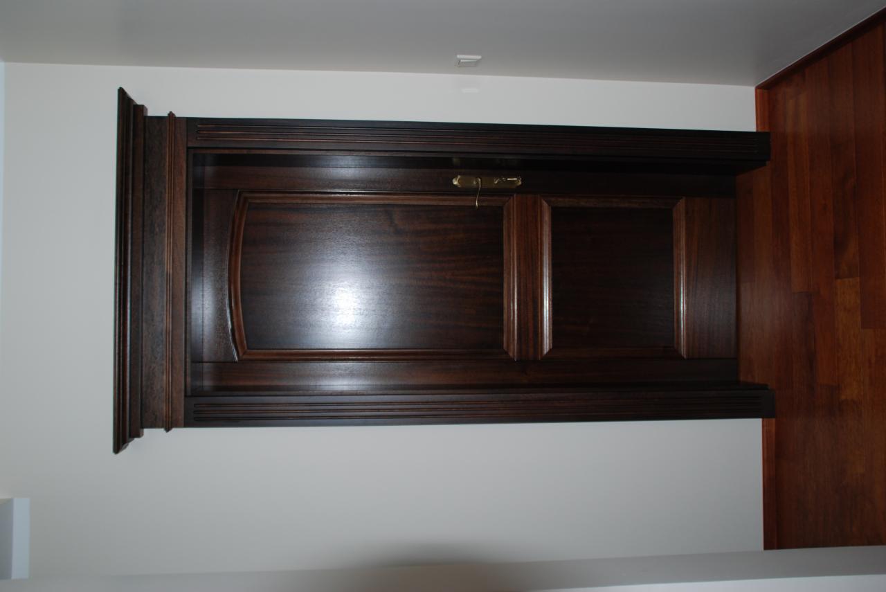 Ciemnobrązowe, drewniane drzwi wewnętrzne z ozdobnymi listwami i mosiężnym zamkiem, widoczne od strony wewnętrznej, zamontowane w białej ścianie, z fragmentem drewnianej podłogi po prawej stronie.