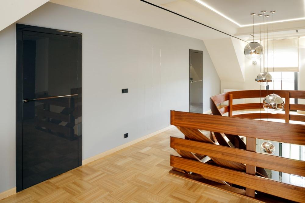 Nowoczesne wnętrze z ciemnymi, lakierowanymi drzwiami z poziomym uchwytem, parkietem w jodełkę i drewnianą balustradą schodów. W tle widoczne kolejne drzwi i designerskie lampy wiszące.