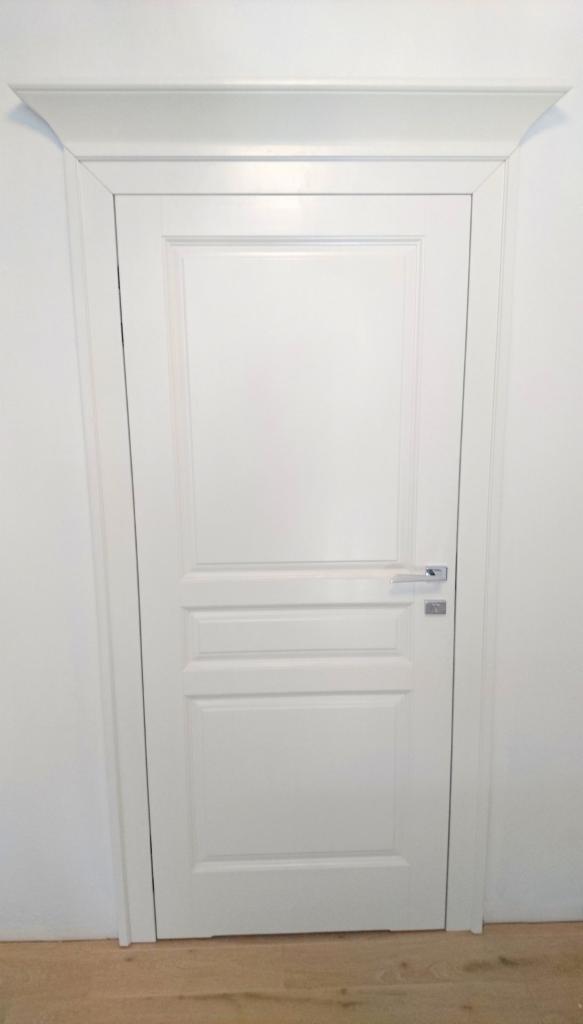 Białe, wewnętrzne drzwi z ozdobną listwą nadprożową, widoczne na tle białej ściany i drewnianej podłogi.