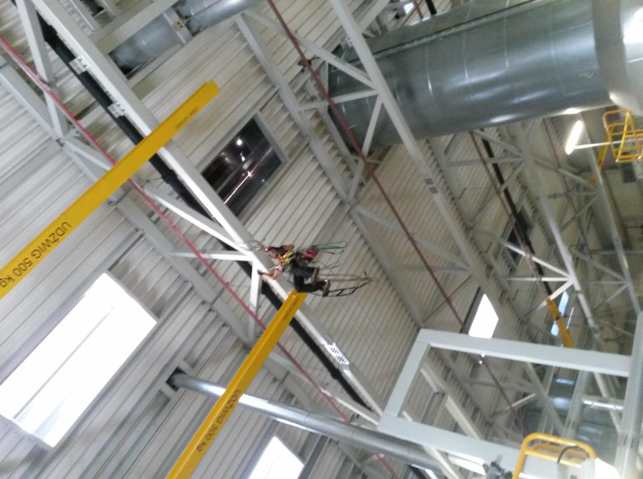 Pracownik na podnośniku koszowym typu nożycowego, wykonujący prace wewnątrz hali produkcyjnej, widoczne stalowe konstrukcje dachu, oświetlenie i instalacje wentylacyjne