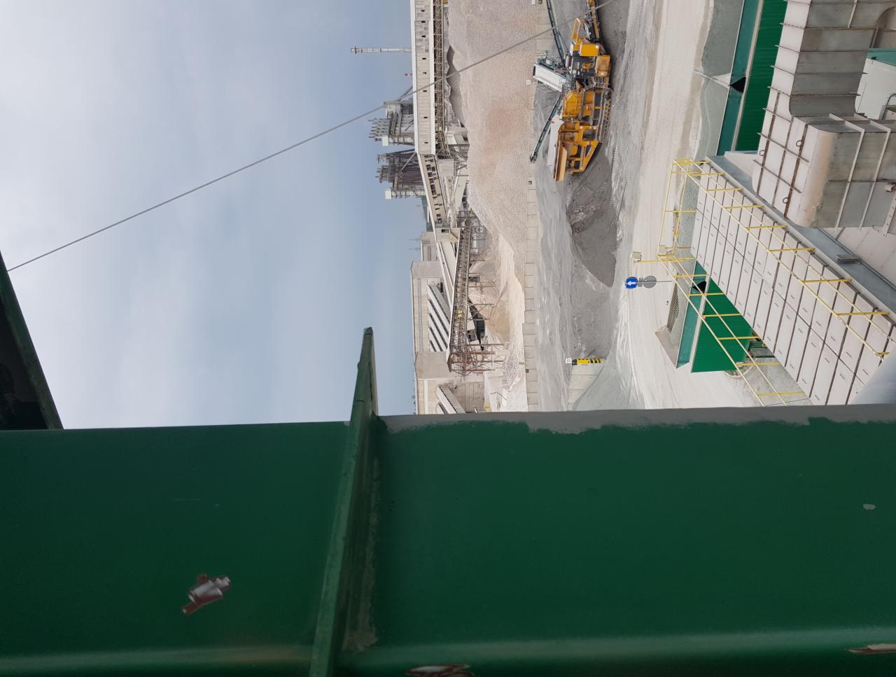 Widok z góry na teren zakładu przemysłowego z żółtą maszyną budowlaną, hałdami piasku i konstrukcjami stalowymi, widziany z zielonej konstrukcji nośnej.