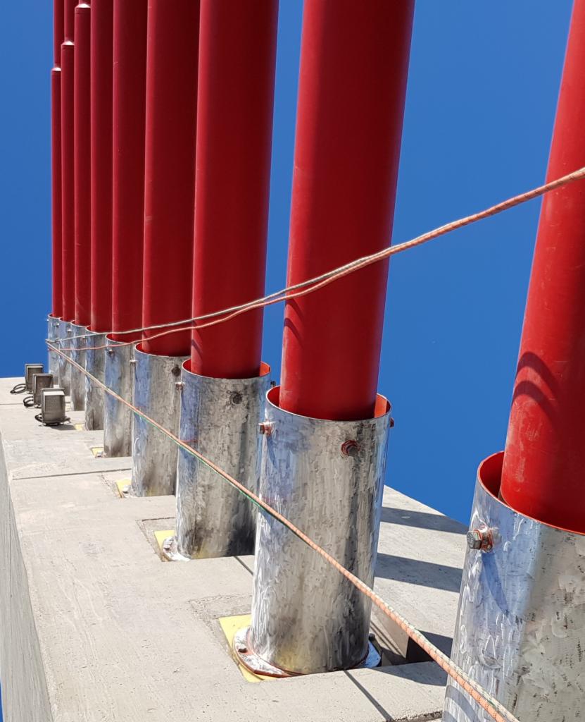 Rząd czerwonych, pionowych rur zabezpieczonych metalowymi osłonami, zamocowanych na betonowej konstrukcji, widoczne liny zabezpieczające na tle błękitnego nieba.