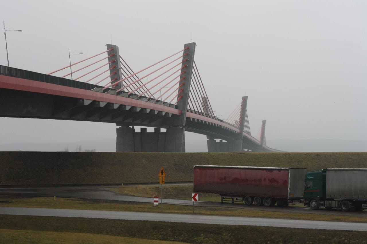 Długi most wantowy z czerwonymi elementami konstrukcyjnymi i betonowymi pylonami w mglisty dzień, z widoczną ciężarówką z naczepą na pierwszym planie.
