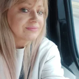 Portret kobiety z blond włosami z różowymi refleksami, w jasnej bluzie, z widocznym oknem pojazdu w tle.