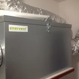 Szara centrala wentylacyjna z widocznymi przewodami wentylacyjnymi w folii aluminiowej, zamontowana przy białej ścianie, z naklejką producenta Enervent.