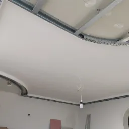 Montaż sufitu podwieszanego z płyt karton-gips na metalowej konstrukcji, widoczne zaokrąglone elementy wykończeniowe, w tle ściana z otworem łukowym i drabina