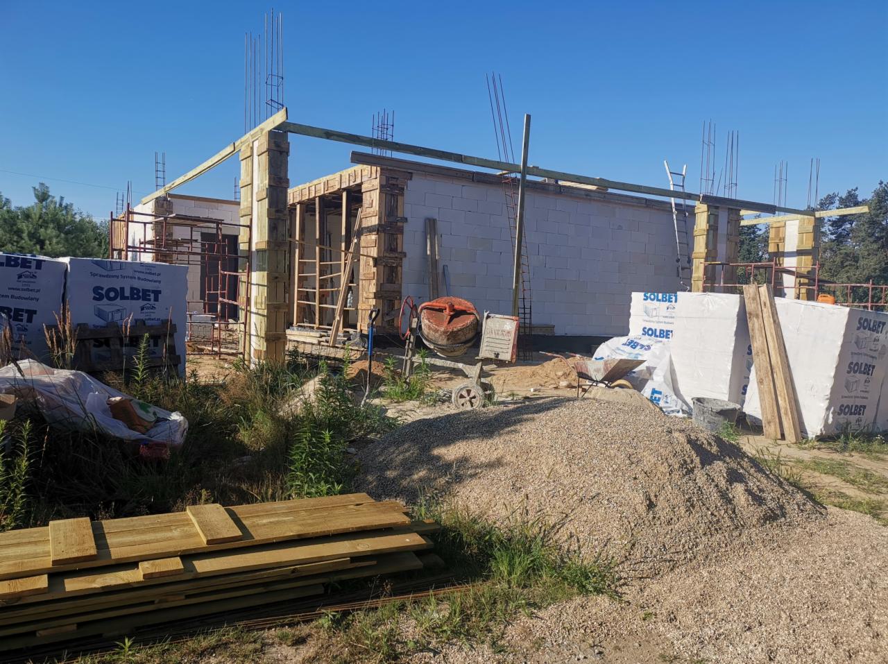 Wznoszenie surowego stanu domu jednorodzinnego z betonu komórkowego Solbet, widoczne zbrojenia, drewniane szalunki, stos materiałów budowlanych i betoniarka.
