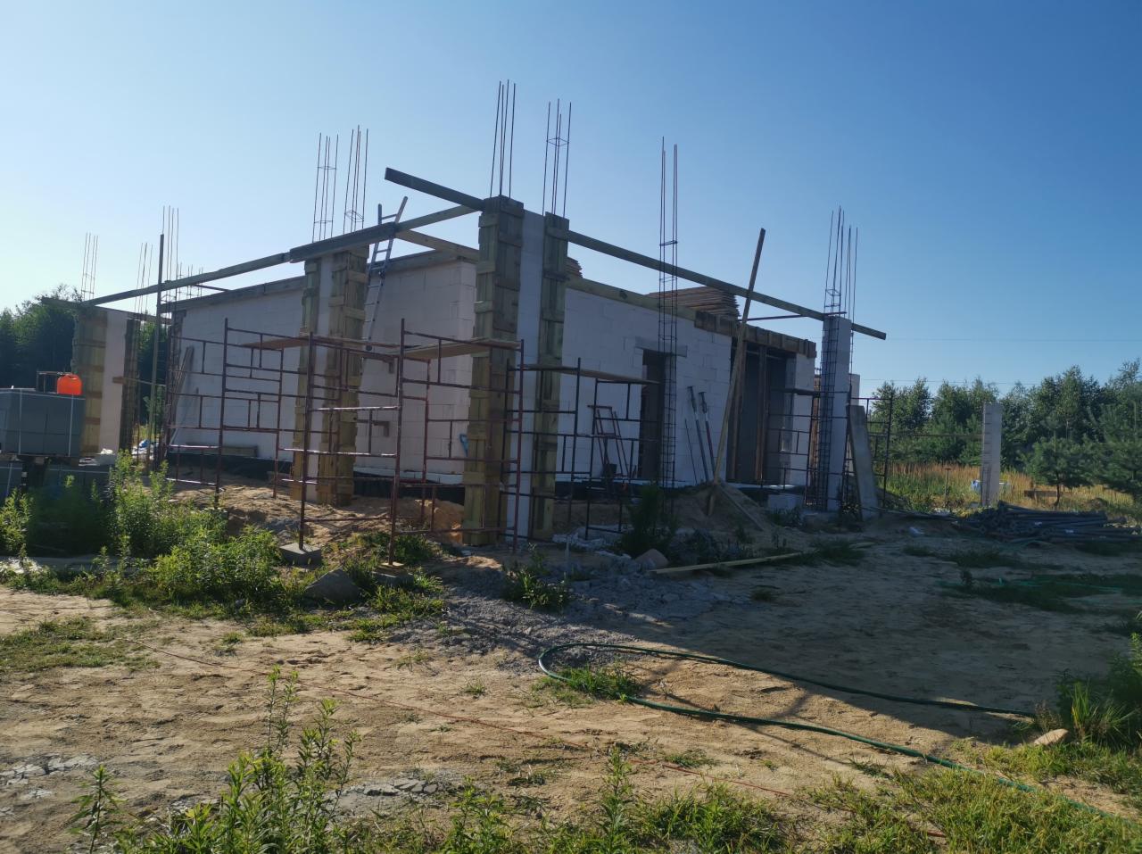 Surowy stan budowy domu jednorodzinnego z białych bloczków, widoczne zbrojenia pionowe i poziome, rusztowania i teren budowy z roślinnością.