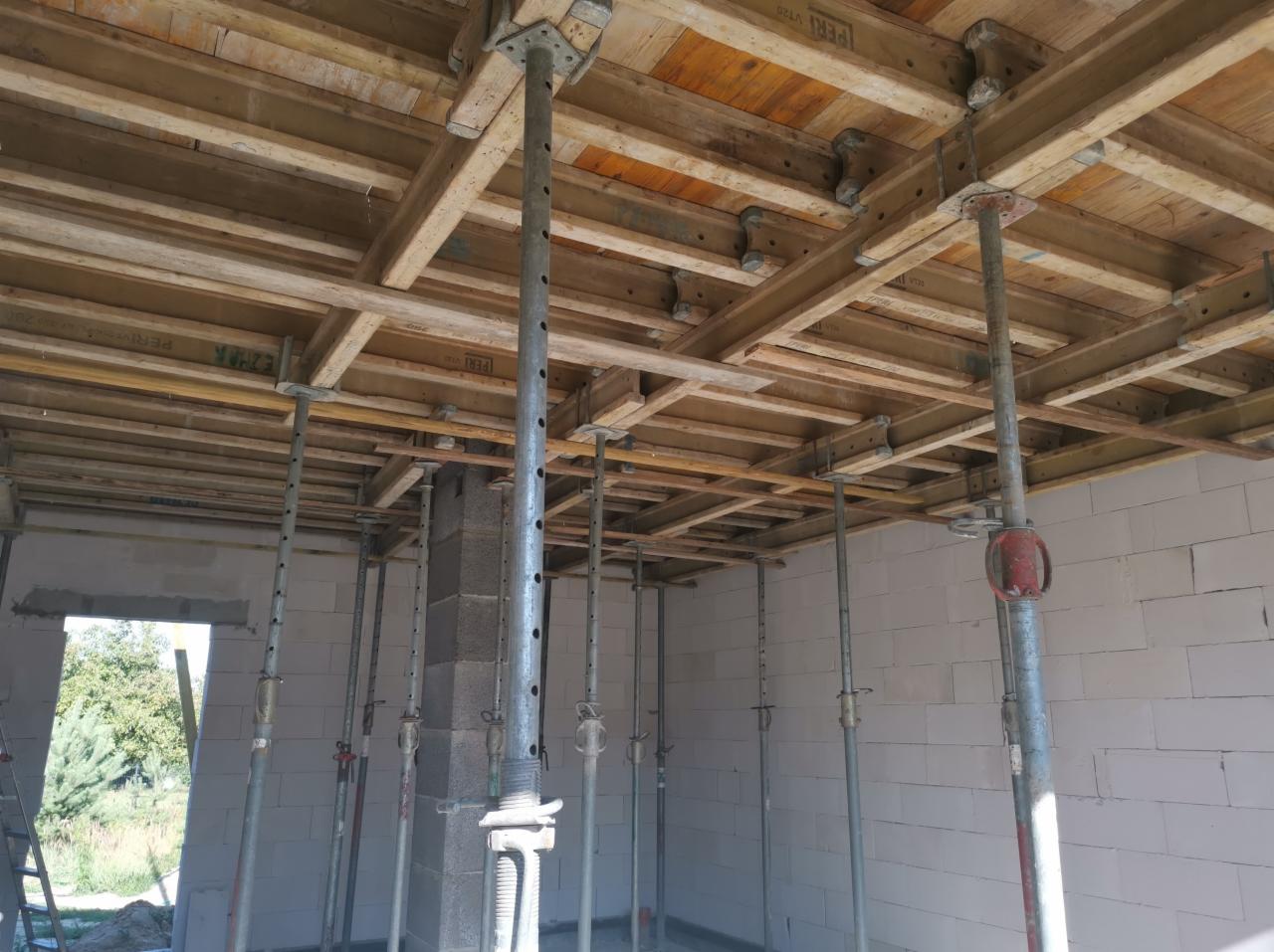 Szare ściany z betonu komórkowego i drewniany szalunek stropowy podparty stalowymi stemplami budowlanymi, widok na zewnątrz przez otwór okienny.